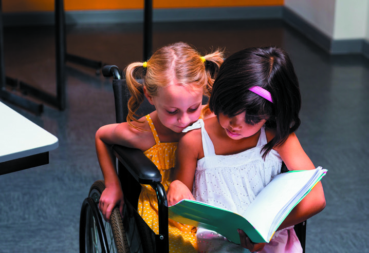 A young girl in wheelchair with another girl sat on her knee both reading a book